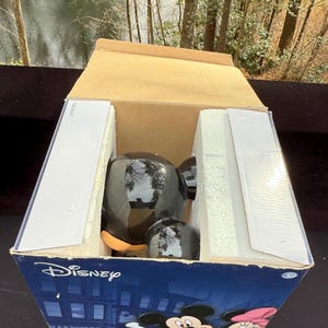 May include: A black Mickey Mouse figurine inside a cardboard box with the Disney logo. The box features illustrations of Mickey and Minnie Mouse. The figurine is glossy and has a round head and ears.