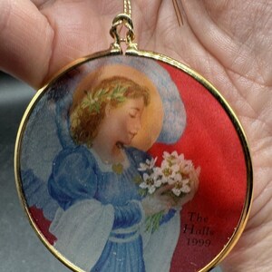 May include: A round, gold-framed ornament with a picture of an angel holding a bouquet of white flowers. The angel is wearing a blue dress and a crown of leaves. The ornament has the text "The Halls 1999" on the bottom.