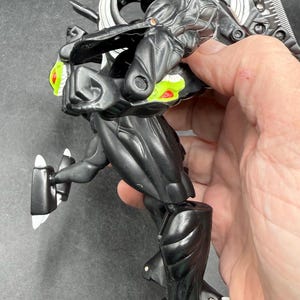 May include: A black and silver action figure with green and red detailing. The figure has a detailed design with articulated joints and a futuristic aesthetic. The figure is held in a hand against a dark background.