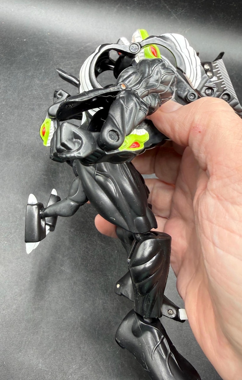 May include: A black and silver action figure with green and red detailing. The figure has a detailed design with articulated joints and a futuristic aesthetic. The figure is held in a hand against a dark background.