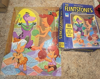 May include: A 100-piece jigsaw puzzle featuring the Flintstones characters. The puzzle depicts Wilma Flintstone, Fred Flintstone, Pebbles Flintstone, and Dino. The puzzle box is blue with a picture of the Flintstones characters on it. The text on the box reads "The Flintstones Jigsaw Puzzle".