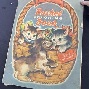 Puede incluir: Libro para colorear "Basket Coloring Book" vintage con una portada azul. La portada muestra una cesta con dos gatitos y un cachorro. El texto dice "Basket Coloring Book" y una etiqueta dice "Carry us home!" en estilo retro.