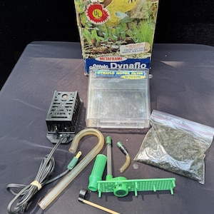 May include: An assortment of aquarium supplies, including a Dynaflo motor filter, tubing, and a bag of gravel. The box features a colorful design with the text "Metaframe Dynaflo". Other components include a black filter, green plastic parts, and a clear plastic container.
