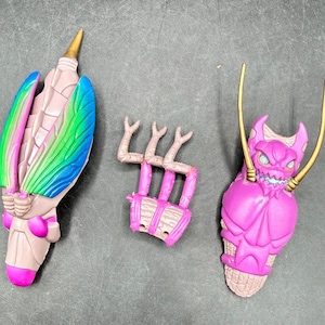 May include: Three colorful toy parts are displayed against a gray background. One is a light brown and pink object with blue, green, and pink wings. Another is a pink, cage-like structure. The third is a pink, cone-shaped object with a monster face.
