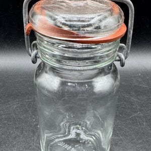 Puede incluir: Tarro de vidrio transparente con tapa abisagrada y asa metálica. La tapa tiene un sello de goma roja. El tarro es cilíndrico con boca ancha y superficie ligeramente texturizada. Diseñado para la conservación de alimentos.