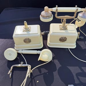 Pode incluir: Telefone antigo creme com mostrador rotativo e detalhes em latão. O telefone possui um auscultador, uma base e duas unidades de base quadradas com um emblema decorativo. O design é reminiscente da tecnologia do início do século XX.