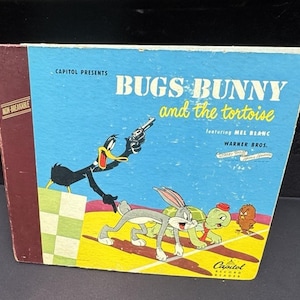 May include: Vintage "Bugs Bunny and the Tortoise" record featuring Mel Blanc. The cover has a blue background with Bugs Bunny, Daffy Duck, a tortoise, and a small bird. The record is labeled "Capitol Record Reader."