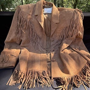 Puede incluir: Una chaqueta de ante color canela con largos flecos en el pecho, hombros, mangas y dobladillo. La chaqueta tiene cuello de solapa, cierre frontal de botones y dos bolsillos. Una etiqueta blanca interior es visible en el cuello.