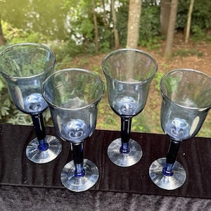 Op de afbeelding: Vier blau getinte wijnglazen met een tulpvormige kelk en een donkerblauwe steel en voet. De glazen zijn in een rij gerangschikt en tonen hun elegante ontwerp en het spel van licht door het glas.