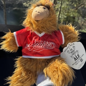 May include: A brown furry puppet wearing a red shirt with the word "Abiter" printed on it. The puppet is holding a tag that says "Collect all 4 of the many faces of ALF. Record & Puppet Sets Only at ALF"