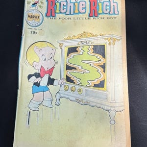 May include: A vintage "Richie Rich" comic book cover. The cover features a cartoon boy in a tuxedo and shorts, looking at a television screen displaying a green dollar sign. The comic book title is in red and blue, with the text "The Poor Little Rich Boy".