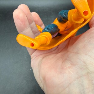 May include: An orange action figure with blue accents on the knees and shoulders. The figure is lying on its back, with its arms and legs extended. The figure has a yellow cape.