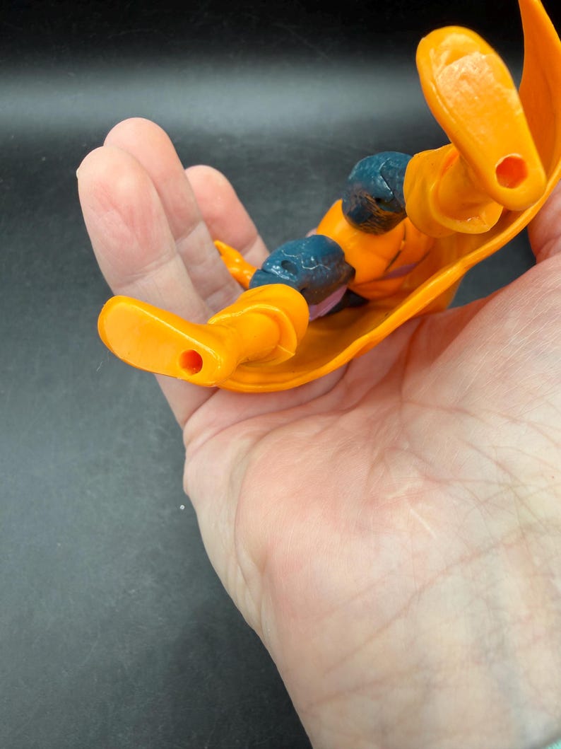 May include: An orange action figure with blue accents on the knees and shoulders. The figure is lying on its back, with its arms and legs extended. The figure has a yellow cape.
