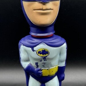 May include: A vintage Batman bobblehead figurine. The figure is primarily blue and white, with a tan face and a yellow belt. The Batman logo is visible on the chest. The figurine is standing on a blue base.