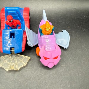 May include: Two toy vehicles featuring Marvel characters. One is a blue and red car with a Spider-Man figure. The other is a pink and purple glider with a Green Goblin figure. A clear plastic spiderweb is also present.