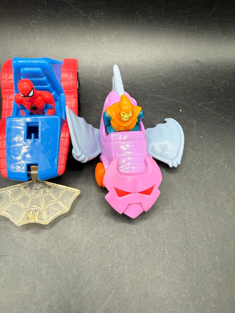 May include: Two toy vehicles featuring Marvel characters. One is a blue and red car with a Spider-Man figure. The other is a pink and purple glider with a Green Goblin figure. A clear plastic spiderweb is also present.