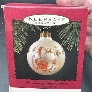 May include: A Keepsake ornament in a red box featuring a white Christmas ball with a Peter Rabbit illustration. The ball has gold accents and the year 1994. The box reads "The Tale of Peter Rabbit" by Beatrix Potter.