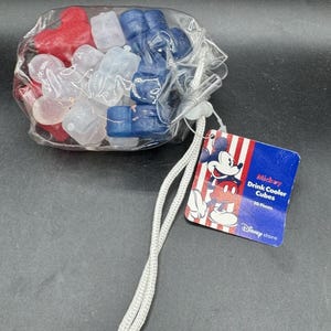 May include: A clear plastic bag filled with red, white, and blue Mickey Mouse-shaped drink cooler cubes. A tag with Mickey Mouse and the text "Mickey Drink Cooler Cubes 20 Pieces" is attached with a white cord.
