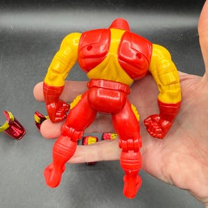 May include: A red and yellow action figure of a superhero, with detachable parts. The figure has a red helmet, chest, arms, and legs, with yellow accents on the shoulders and arms. The figure is in a dynamic pose.