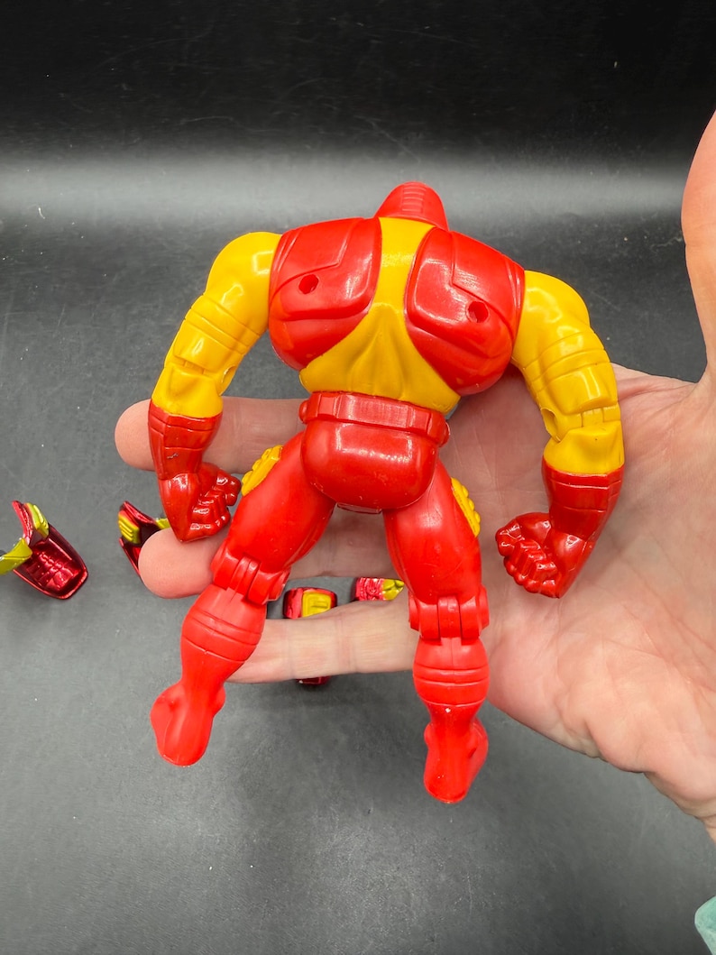 May include: A red and yellow action figure of a superhero, with detachable parts. The figure has a red helmet, chest, arms, and legs, with yellow accents on the shoulders and arms. The figure is in a dynamic pose.