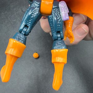 May include: A close-up of a toy action figure with orange and blue parts. The figure has orange boots, a pumpkin-shaped torso, and a matching orange cape. The figure's legs are blue with a textured pattern. A small orange ball is next to the figure.