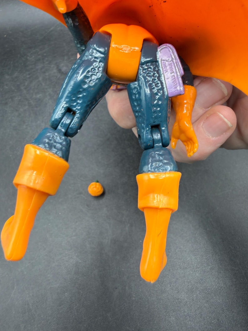 May include: A close-up of a toy action figure with orange and blue parts. The figure has orange boots, a pumpkin-shaped torso, and a matching orange cape. The figure's legs are blue with a textured pattern. A small orange ball is next to the figure.