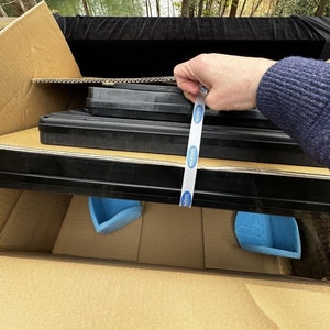 Op de afbeelding: Een kartonnen doos met een zwart en grijs item erin. Het item is verpakt in blauw schuim en vastgemaakt met een witte tape met het woord "Intertape" erop gedrukt.