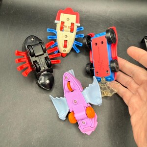 May include: Four colourful toy cars, including a red and blue car, a black car, a pink bat-shaped car, and a white car. The cars have wheels and are designed with insect-like and bat-like features.