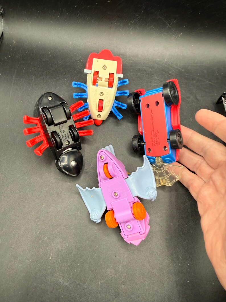 May include: Four colourful toy cars, including a red and blue car, a black car, a pink bat-shaped car, and a white car. The cars have wheels and are designed with insect-like and bat-like features.