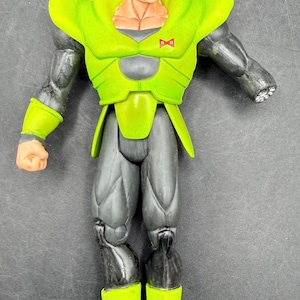 May include: A detailed action figure of Android 16 from the Dragon Ball Z series. The figure features a green and black color scheme, with a distinctive orange hairstyle. The figure is posed with arms at its sides, showcasing its muscular build.