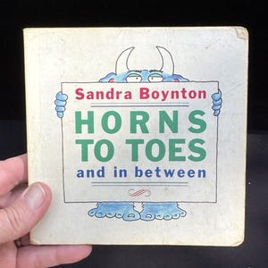 May include: A square, off-white children's book titled "Horns to Toes and in between" by Sandra Boynton. The cover features a blue monster holding a sign with the title in green and the author's name in pink.