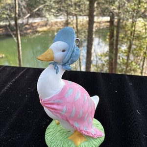 Pode incluir: Uma figurinha de porcelana branca de um ganso usando um chapéu azul e um xale rosa com uma base verde. O ganso está em pé sobre uma base verde com um bico e pés amarelos.