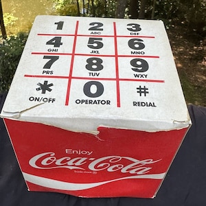May include: A vintage Coca-Cola cardboard box with a red and white design. The box features a classic Coca-Cola logo and a vintage rotary phone dial pad with numbers and letters. The text "Enjoy Coca-Cola" and "Trade-mark" are printed on the box.