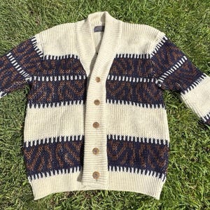 May include: Cream-coloured cardigan with a shawl collar and button closure. The sweater features horizontal stripes in navy blue and brown, with a textured knit design. The sleeves and body have a relaxed fit.