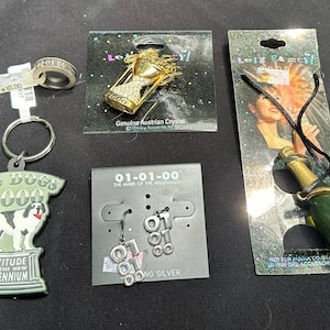 Peut inclure: Assortiment d'objets fantaisie sur le thème du millénaire, comprenant un porte-clés avec un chien, une bague, une épingle avec un sablier incrusté de cristaux, des boucles d'oreilles et un collier avec un pendentif en forme de bouteille de champagne. Les objets sont présentés sur une surface sombre.