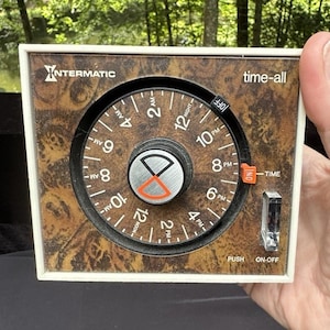 May include: A square Intermatic Time-All electrical timer with a faux wood grain faceplate. The circular dial features white numbers for 12-hour time, an orange indicator, and a central silver knob. A clear plastic push switch is labeled ON-OFF.