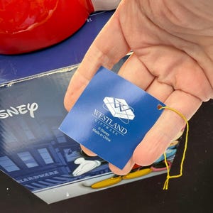 May include: A blue tag with the Westland Giftware logo and Disney branding is held in front of a red object and a box with the Disney logo. The tag has a gold string attached. The box has a Mickey Mouse design.
