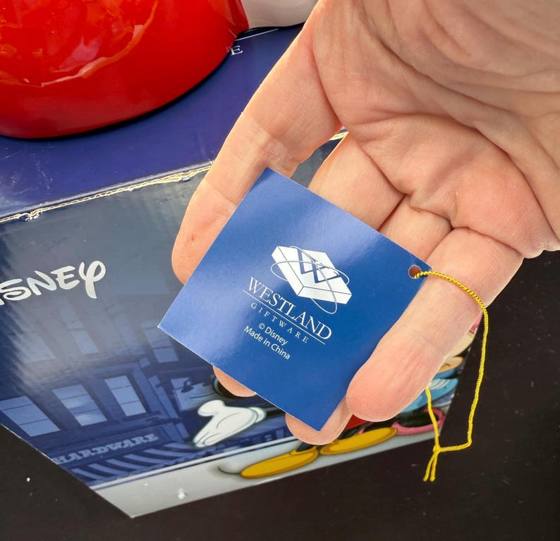 May include: A blue tag with the Westland Giftware logo and Disney branding is held in front of a red object and a box with the Disney logo. The tag has a gold string attached. The box has a Mickey Mouse design.
