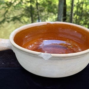 May include: A cream-coloured ceramic bowl with a handle, featuring a rich brown interior. The bowl has a slightly chipped rim. The background is blurred, showing green foliage.