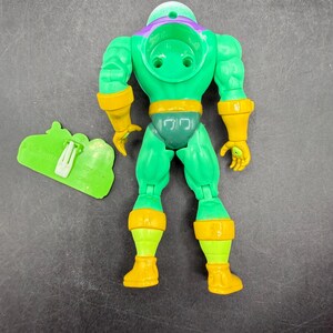 Op de afbeelding: Een groene actiefiguur met gele en paarse accenten. De figuur heeft een helm, handschoenen en laarzen. Een groen accessoire met de tekst "TOY HITS" staat naast de figuur.