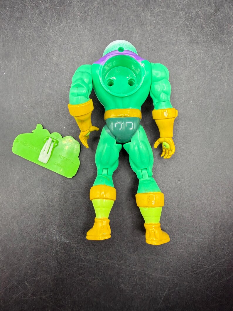 Op de afbeelding: Een groene actiefiguur met gele en paarse accenten. De figuur heeft een helm, handschoenen en laarzen. Een groen accessoire met de tekst "TOY HITS" staat naast de figuur.