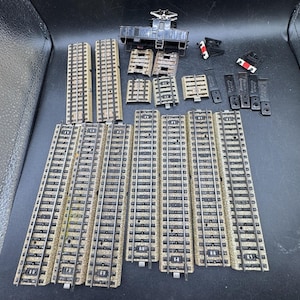 Puede incluir: Un conjunto de piezas de vías de tren en miniatura, que incluye vías rectas, vías curvas y un vagón. Las vías son de color beige y marrón, con raíles plateados. Accesorios: topes y un vagón negro.