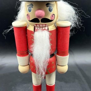 Könnte beinhalten: Ein traditioneller Nussknacker aus Holz mit einer rot-goldenen Uniform, einem weißen Bart und Schnurrbart und einem schwarzen Hut. Der Nussknacker steht auf einem roten Sockel.