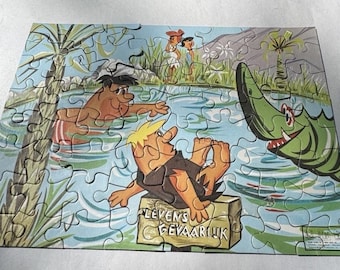 May include: A vintage jigsaw puzzle depicting a cartoon scene of three cavemen in a jungle setting. One caveman is swimming in a lake, another is sitting on a log, and the third is standing on a rock. The puzzle is made of cardboard and has a blue background with green foliage. The puzzle is titled "Levens Gevaarlijk" which translates to "Life Dangerous".