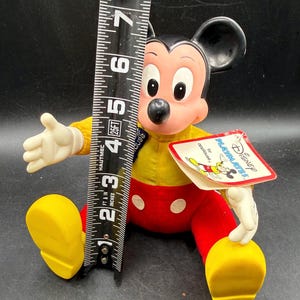 May include: A vintage Mickey Mouse plush doll, approximately 6 inches tall, wearing a yellow shirt, red pants with white buttons, and yellow shoes. The doll has a tag that reads "Disney PLAYMATES by applause."