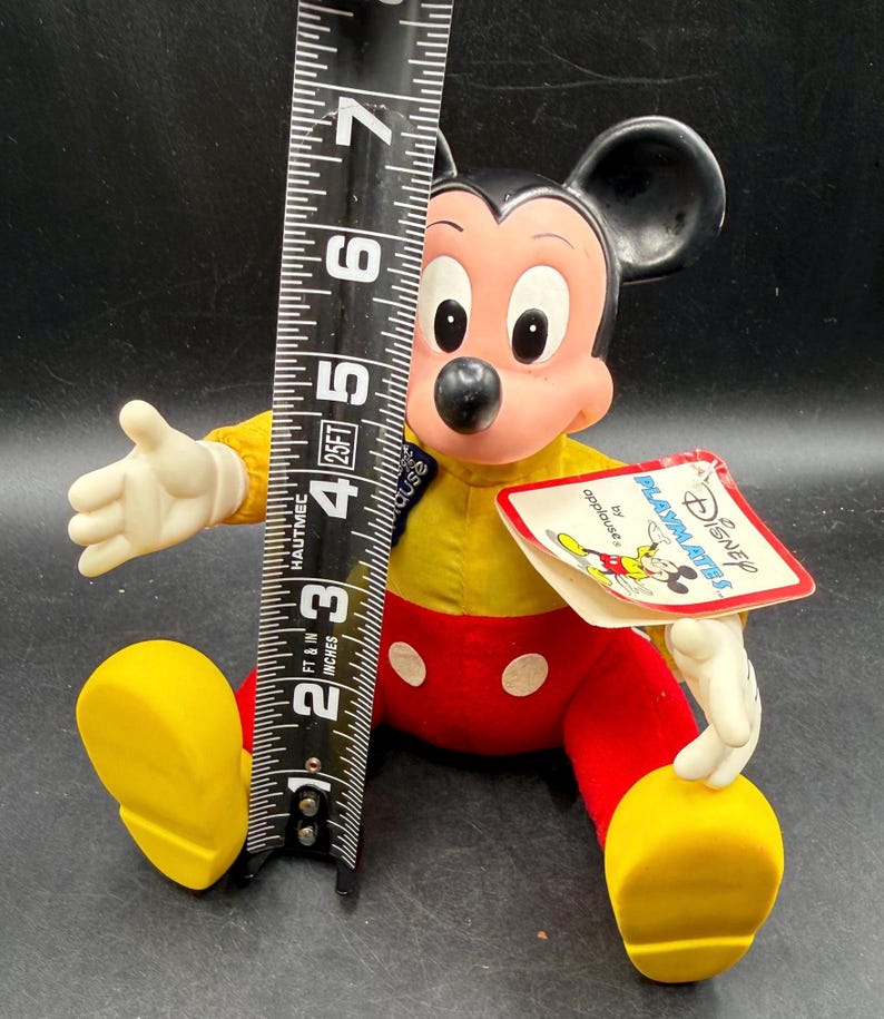 May include: A vintage Mickey Mouse plush doll, approximately 6 inches tall, wearing a yellow shirt, red pants with white buttons, and yellow shoes. The doll has a tag that reads "Disney PLAYMATES by applause."