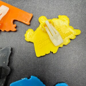 Może przedstawiać: Kolekcja kolorowych plastikowych klips&oacute;w do zabawek w kolorach pomarańczowym, ż&oacute;łtym, niebieskim i szarym. Klipsy mają r&oacute;żne kształty i zawierają tekst, np. "MARVEL &copy;1995" i "TOY BIZ &copy;1994". Klipsy są wyprodukowane w Chinach.