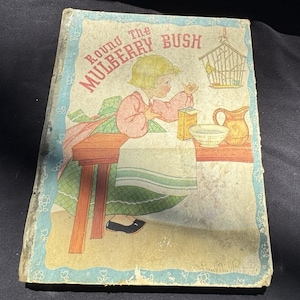 May include: Vintage children's book titled "Round The Mulberry Bush." The cover features a colorful illustration of a young girl with blonde hair, a birdcage, and various kitchen items. The book has a light blue border with floral accents.