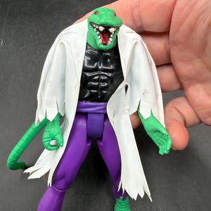 K&ouml;nnte beinhalten: Eine gr&uuml;ne Echsenkopf-Actionfigur mit wei&szlig;em Laborkittel, schwarzem Torso, lila Hose und gr&uuml;nen H&auml;nden und F&uuml;&szlig;en. Die Figur hat einen offenen Mund mit wei&szlig;en Z&auml;hnen und einer roten Zunge. Die Figur wird von einer Hand gehalten.
