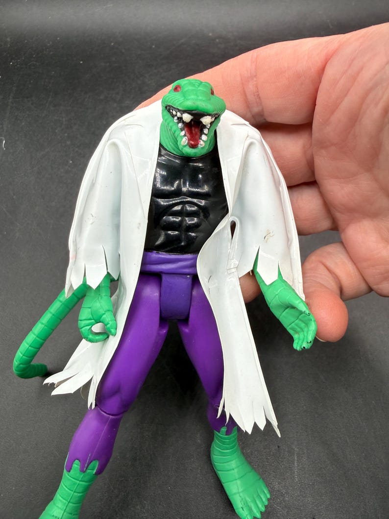 K&ouml;nnte beinhalten: Eine gr&uuml;ne Echsenkopf-Actionfigur mit wei&szlig;em Laborkittel, schwarzem Torso, lila Hose und gr&uuml;nen H&auml;nden und F&uuml;&szlig;en. Die Figur hat einen offenen Mund mit wei&szlig;en Z&auml;hnen und einer roten Zunge. Die Figur wird von einer Hand gehalten.